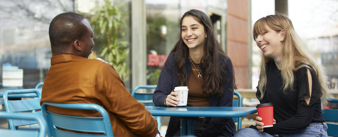 three students sat in blue metal chairs at a blue outdoor table, two are holding takeaway coffee cups